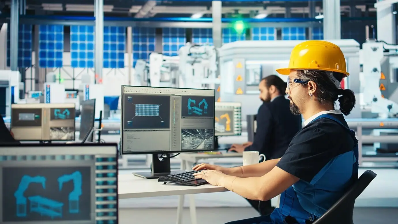 A photovoltaics factory engineer in a yellow hard hat analyzing data on a PC.